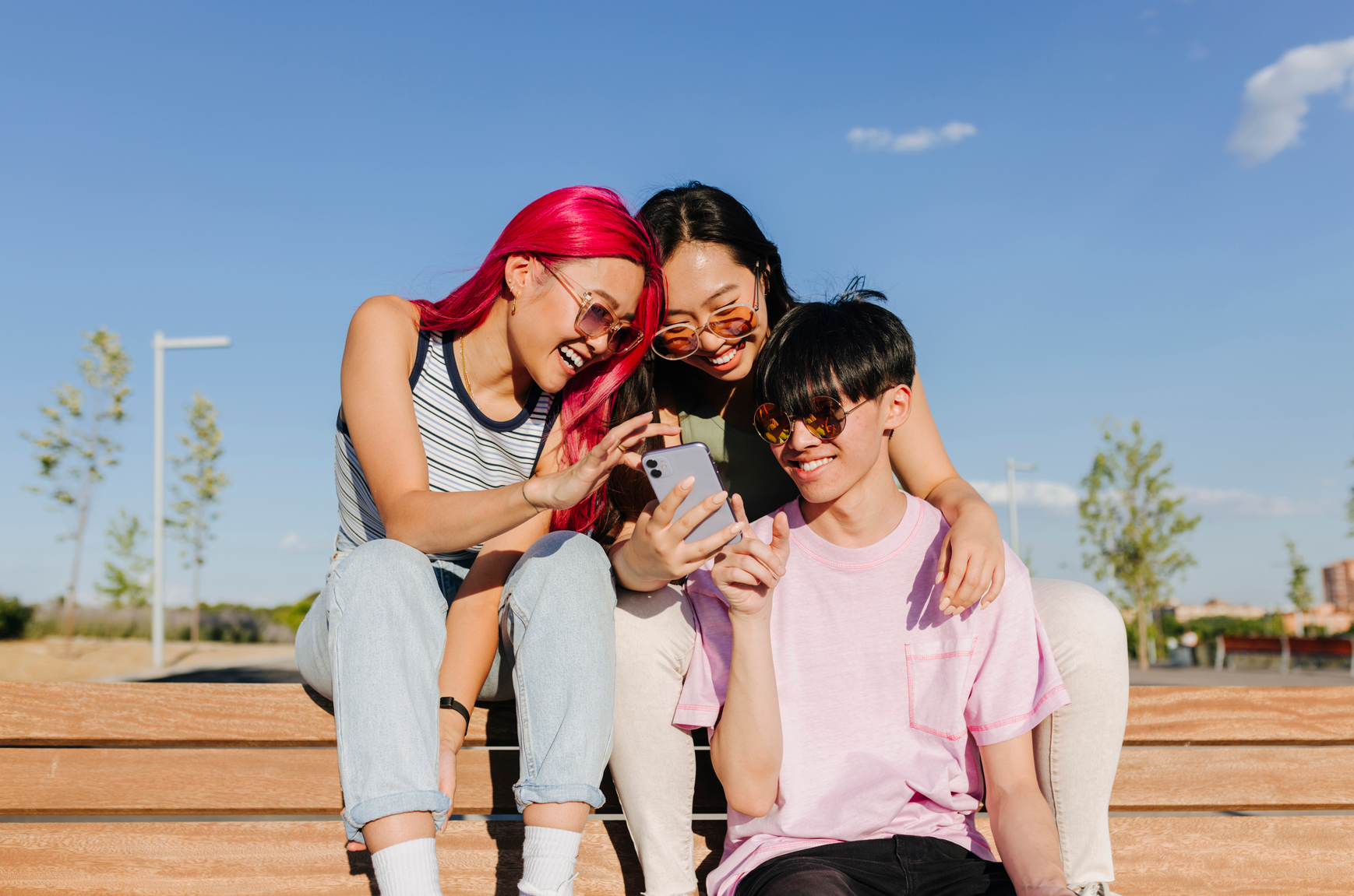 Positive young friends in casual clothes sitting on wooden bench checking social networks on mobile phone